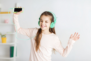 people, children, pajama party and technology concept - happy smiling girl in headphones with smartphone and listening to music at home