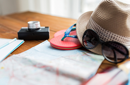 summer vacation, tourism and objects concept - close up of travel map, flip-flops, hat and camera on wooden table at home