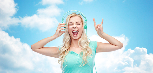 music, technology and people concept - happy young woman or teenage girl with headphones singing song and showing rock gesture over blue sky and clouds background