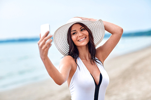 lifestyle, leisure, summer, technology and people concept - smiling young woman or teenage girl in sun hat taking selfie with smartphone on beach