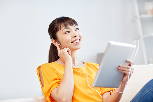 people, technology and leisure concept - happy asian young woman sitting on sofa with tablet pc computer and earphones listening to music at home