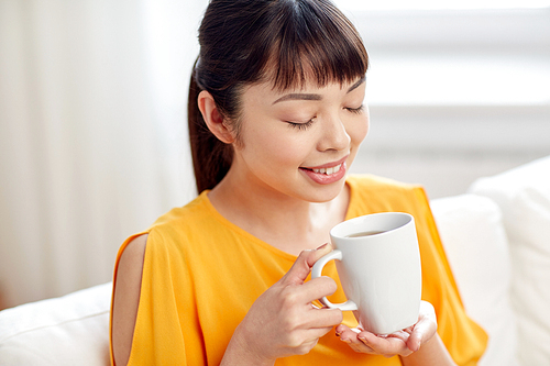 people, drinks and leisure concept - happy asian woman sitting on sofa and drinking tea from cup or mug at home