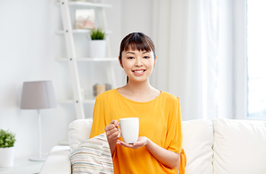 people, drinks and leisure concept - happy asian woman sitting on sofa and drinking tea from cup or mug at home