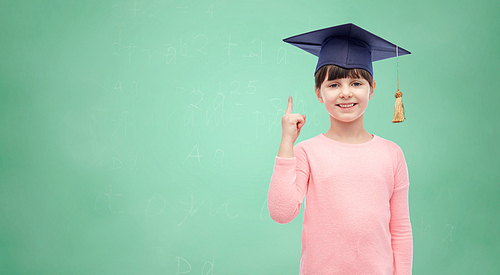 childhood, school, education, learning and people concept - happy girl with in bachelor hat or mortarboard over green school chalk board background