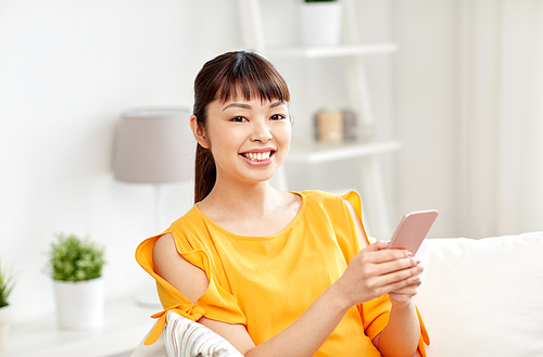 people, technology, communication and leisure concept - happy young asian woman sitting on sofa and texting message on smartphone at home