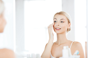 beauty, skin care and people concept - smiling young woman applying cream to face and looking to mirror at home bathroom