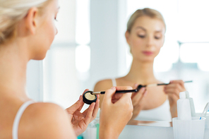 beauty, make up, cosmetics, morning and people concept - close up of young woman applying eyeshade with makeup brush and looking to mirror at home bathroom