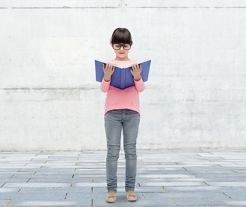 childhood, school, education, vision and people concept - happy little girl in eyeglasses reading book over urban city street background