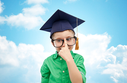 childhood, school, education, knowledge and people concept - happy boy in bachelor hat or mortarboard and eyeglasses over blue sky and clouds background