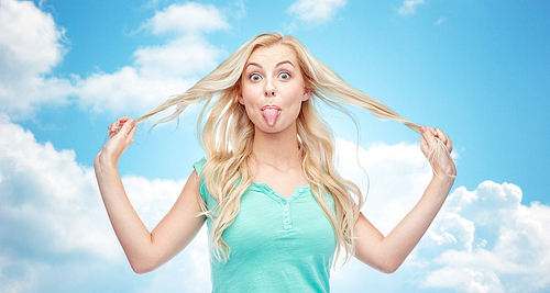 emotions, expressions, hairstyle and people concept - smiling young woman or teenage girl showing her tongue and holding strand of hair over blue sky and clouds background