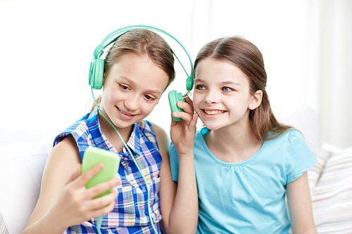 people, children, technology, friends and friendship concept - happy little girls with smartphone and earphones sitting on sofa and listening to music at home