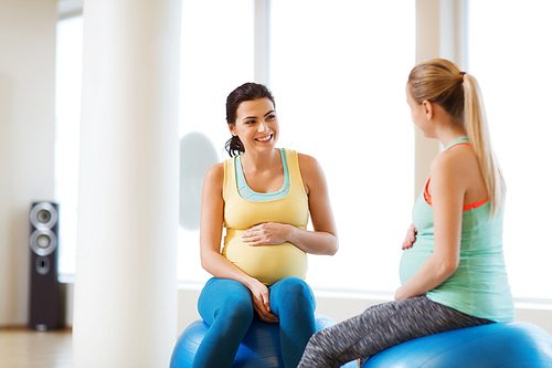 pregnancy, sport, fitness, people and healthy lifestyle concept - two happy pregnant women sitting and talking on balls in gym