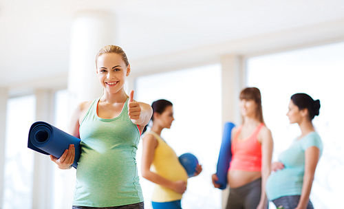 pregnancy, sport, fitness, people and healthy lifestyle concept - happy pregnant woman with mat in gym showing thumbs up