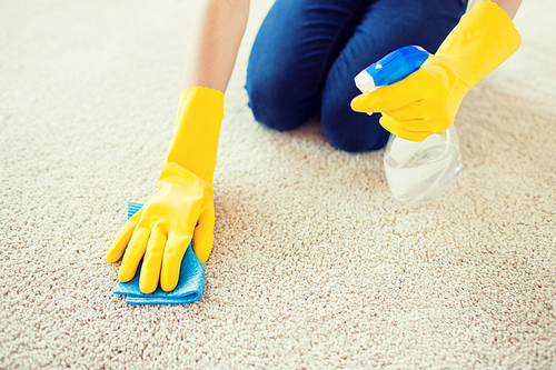 people, housework and housekeeping concept - close up of woman in rubber gloves with cloth and detergent spray cleaning carpet at home