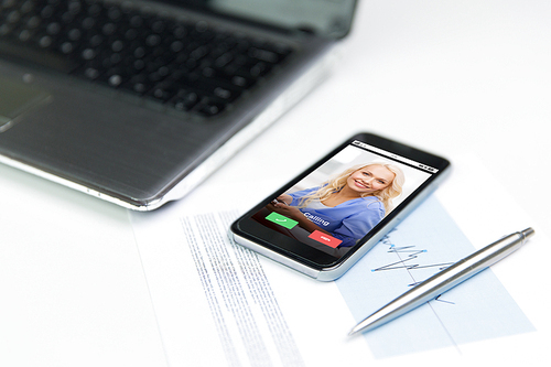 business, technology and statistics concept - close up of smartphone with incoming call on screen, laptop computer and pen on office table