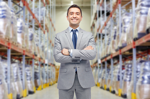 wholesale, logistic, business, export and people concept - happy man in suit and tie at warehouse over warehouse background