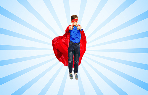 happiness, freedom, childhood, movement and people concept - boy in red super hero cape and mask flying in air and showing thumbs up over blue burst rays background