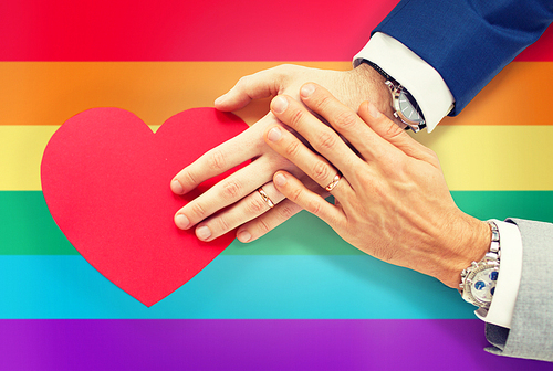 people, homosexuality, same-sex marriage, valentines day and love concept - close up of happy married male gay couple hands with red paper heart shape over rainbow flag background