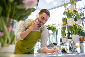 people, sale, retail, business and floristry concept - happy smiling florist man calling on smartphone and making notes to clipboard at flower shop counter