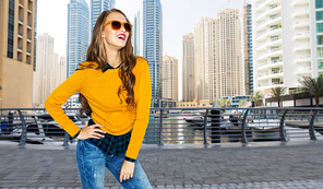 people, travel, tourism, style and fashion concept - happy young woman or teen girl in casual clothes and sunglasses over dubai city street background
