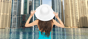 people, summer holidays, travel, tourism and vacation concept - woman in swimsuit and sun hat from back over dubai city and infinity edge pool background