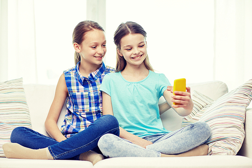 people, children, technology, friends and friendship concept - happy little girls sitting on sofa and taking selfie with smartphone at home