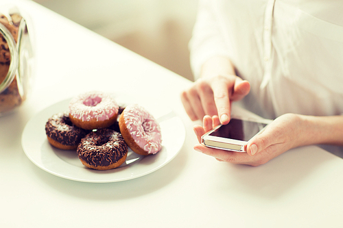 people, junk food, , technology and unhealthy eating concept - close up of hands with smart phone and donuts counting calories and sitting at table at home