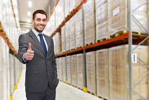 wholesale, logistic, business, export and people concept - happy man in suit and tie showing thumbs up gesture over warehouse background