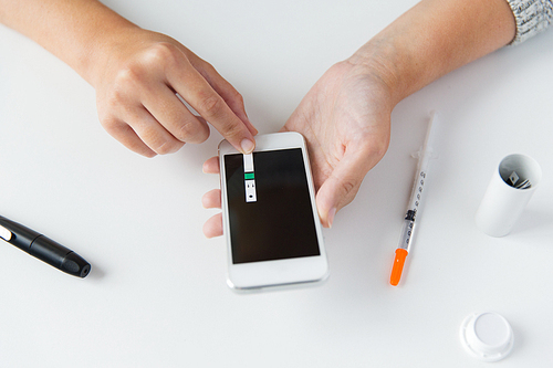 medicine, diabetes, glycemia, health care and people concept - close up of woman hands with smartphone checking blood sugar level by glucometer at home