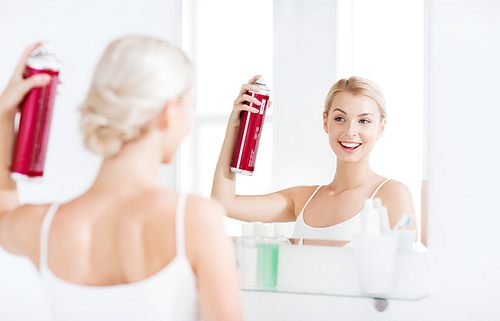 beauty, hygiene, hairstyle, morning and people concept - smiling young woman with hairspray styling her hair and looking to mirror at home bathroom