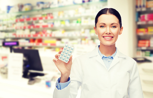 medicine, pharmacy, people, health care and pharmacology concept - happy young woman pharmacist with pills over drugstore background