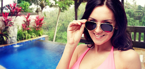 people, summer holidays, travel and tourism concept - happy young woman in sunglasses and pink swimsuit looking at you over hotel resort background