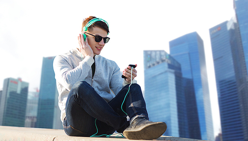 technology, travel, tourism and people concept - smiling young man or teenage boy in headphones with smartphone listening to music over singapore city skyscrapers background