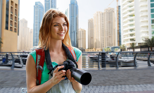 adventure, travel, tourism, hike and people concept - happy young woman with backpack and camera photographing over dubai city and harbor with boats background