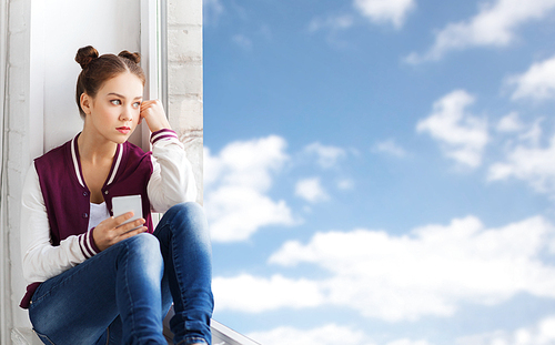 people, emotion, technology and teens concept - sad unhappy pretty teenage girl sitting on windowsill with smartphone and looking through window over blue sky and clouds background