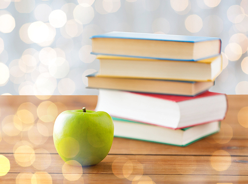 education, school, literature, reading and knowledge concept - close up of books and green apple on wooden table over holidays lights background