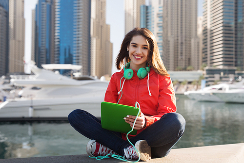 technology, travel, tourism, music and people concept - smiling young woman or teenage girl with tablet pc computer and headphones over dubai city street and boats in harbor background