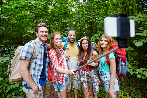 technology, travel, tourism, hike and people concept - group of smiling friends walking with backpacks taking picture by smartphone on selfie stick in woods