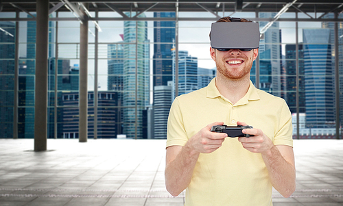 3d technology, virtual reality, entertainment and people concept - happy young man with virtual reality headset playing with game controller gamepad over empty industrial room and city background