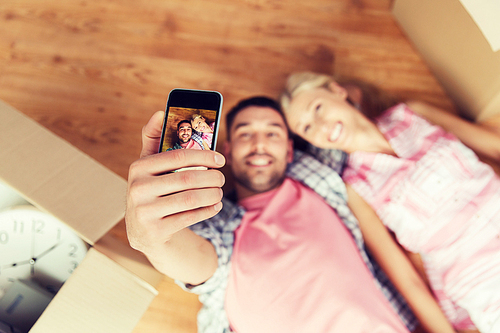 new home, technology, people, repair and moving concept - happy couple taking selfie with smartphone and lying on floor among cardboard boxes at home