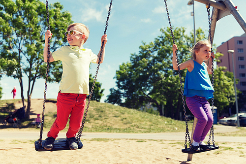 summer, childhood, leisure, friendship and people concept - two happy kids swinging on swing at children playground