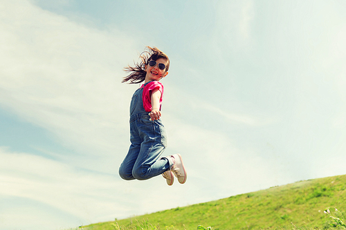 summer, childhood, leisure and people concept - happy little girl jumping high over green field and blue sky outdoors