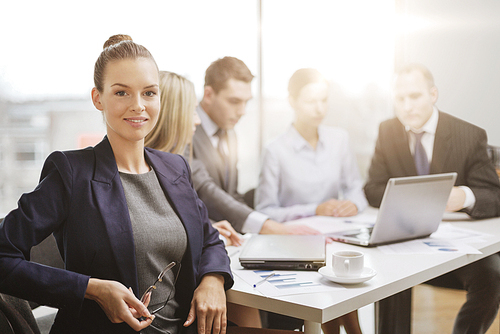 business, technology and office concept - smiling businesswoman with eyeglasses in office with team on the back