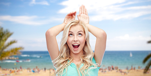 fun, expressions, summer holidays, travel and people concept - happy smiling young woman making bunny ears over exotic tropical beach with palm trees and sea background