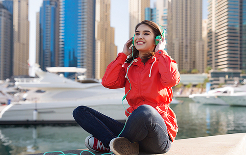 technology, travel, tourism and people concept - smiling young woman or teenage girl in headphones listening to music over dubai city street and boats in harbor background