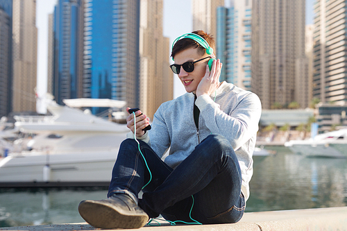 technology, travel, tourism and people concept - smiling young man or teenage boy in headphones with smartphone listening to music over dubai city street and boats in harbor background