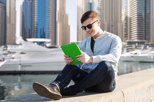 technology, travel, tourism and people concept - smiling young man or teenage boy with tablet pc computers over dubai city street and boats in harbor background