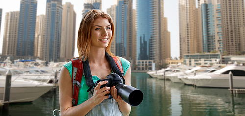 adventure, travel, tourism, hike and people concept - happy young woman with backpack and camera photographing over dubai city and harbor with boats background