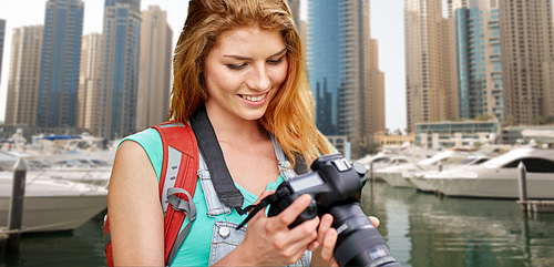 adventure, travel, tourism, hike and people concept - happy young woman with backpack and camera photographing over dubai city and harbor with boats background