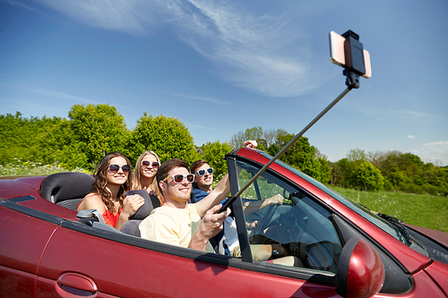 leisure, road trip, travel and people concept - happy friends driving in cabriolet taking picture by smartphone selfie stick at country